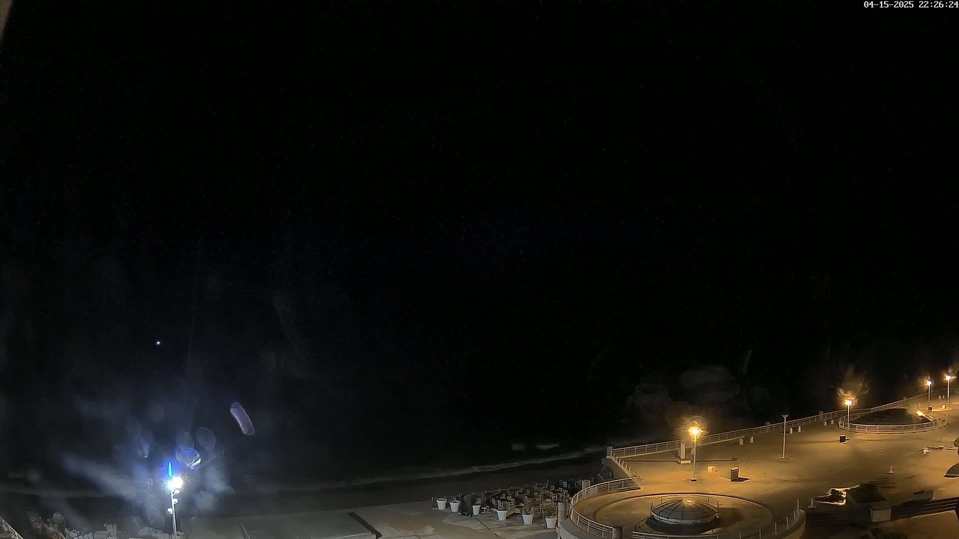 A nighttime view of a mostly empty beachside promenade with waves breaking on the shore under a clear, dark sky.