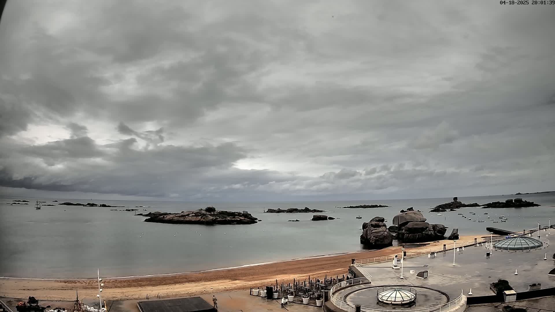 A sandy beach and calm ocean are partially visible beneath an overcast sky, with rocky outcroppings in the water and a paved area with structures in the foreground.