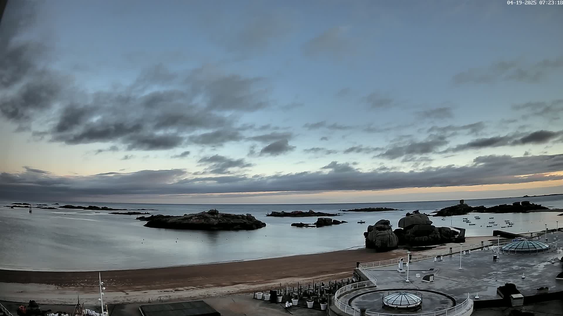 A calm bay with rocky outcrops and a sandy beach is visible under a mostly cloudy sky at dawn.