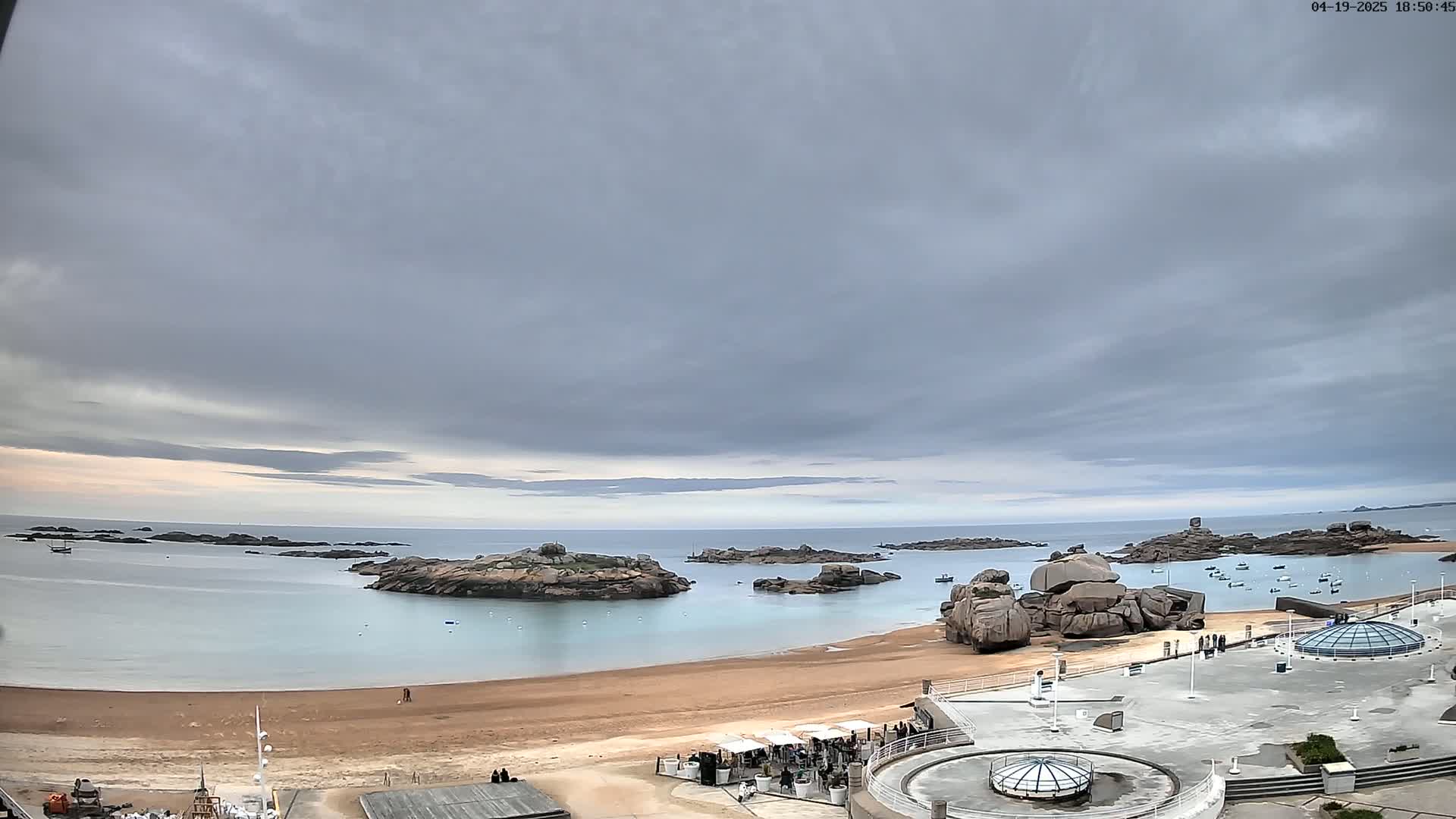 A sandy beach with calm, light-blue water, several rocky outcrops, and a few boats is visible under an overcast sky.