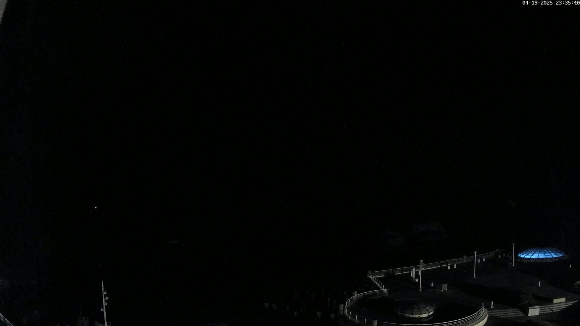 A nighttime, high-angle view shows a dark, empty plaza with a few illuminated structures and light poles.