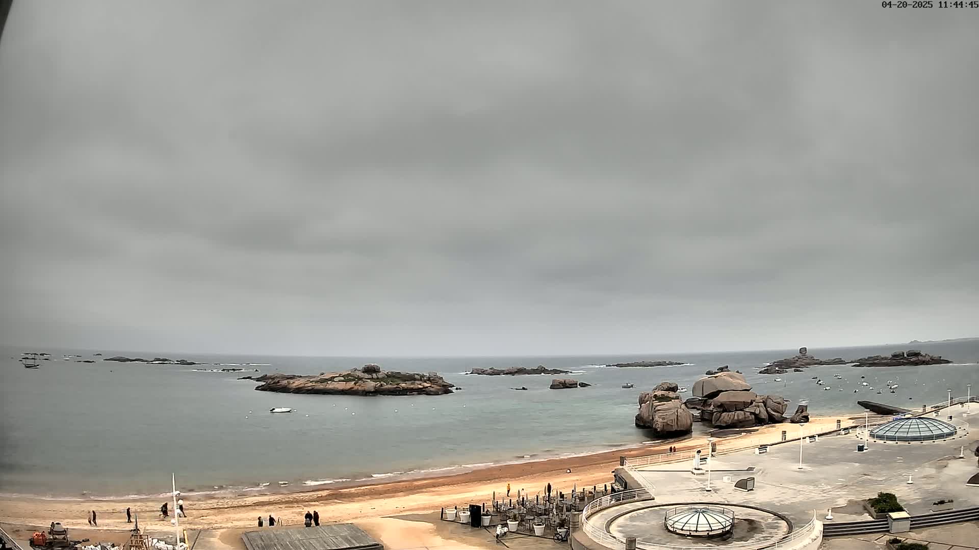 A sandy beach and calm, gray-sky ocean are seen from an elevated vantage point, with several rocky islands and outcroppings in the water and some people on the beach.