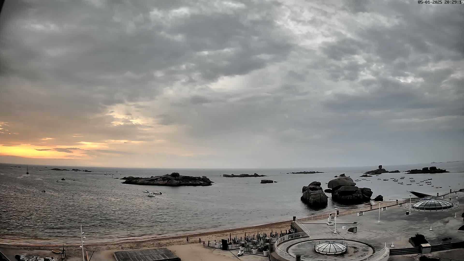 A mostly cloudy sunset over a calm body of water with a sandy beach, several boats, and large rocks near the shore.