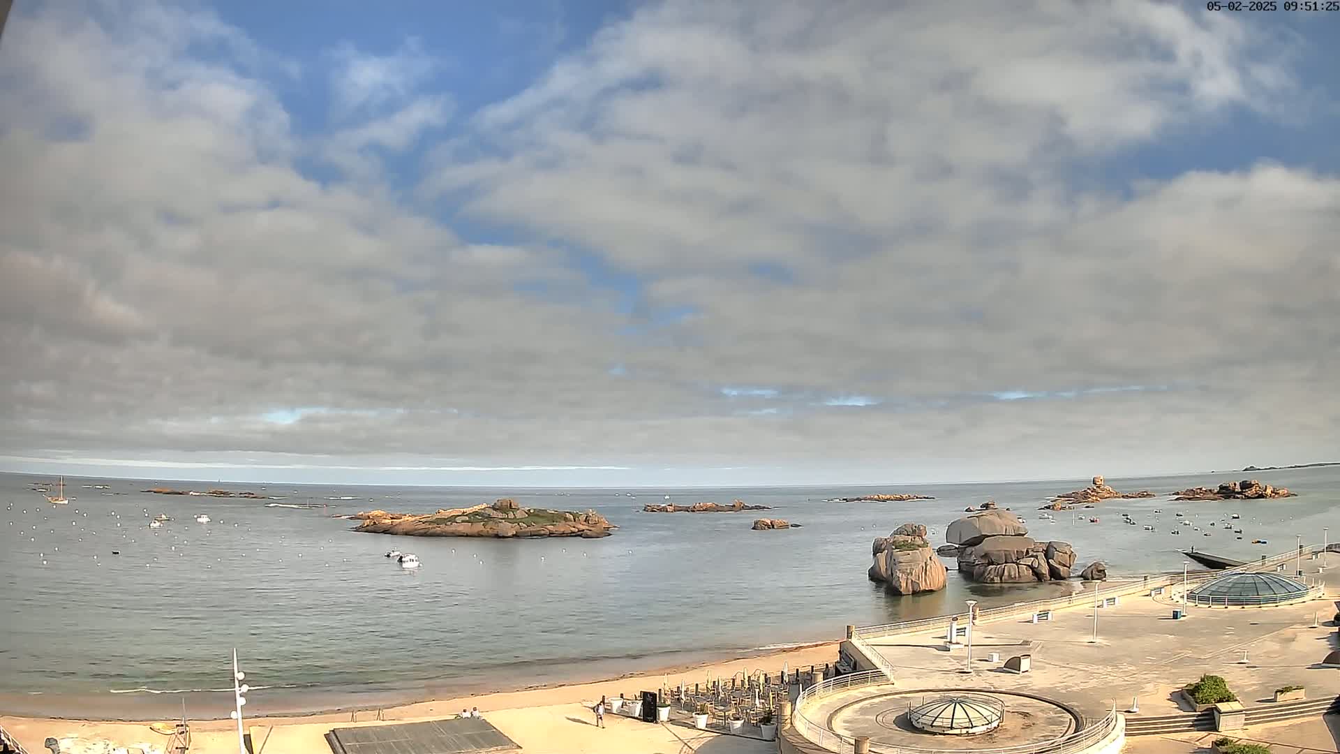 A partly cloudy day reveals a sandy beach, calm ocean water dotted with small boats, and rocky outcrops near a paved waterfront area with circular structures.