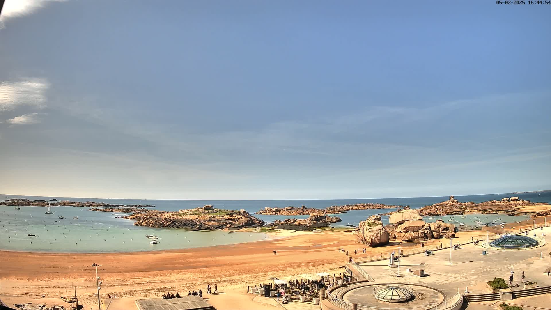 A sandy beach with calm, light-green water is surrounded by pink granite rocks under a mostly clear, light-blue sky.