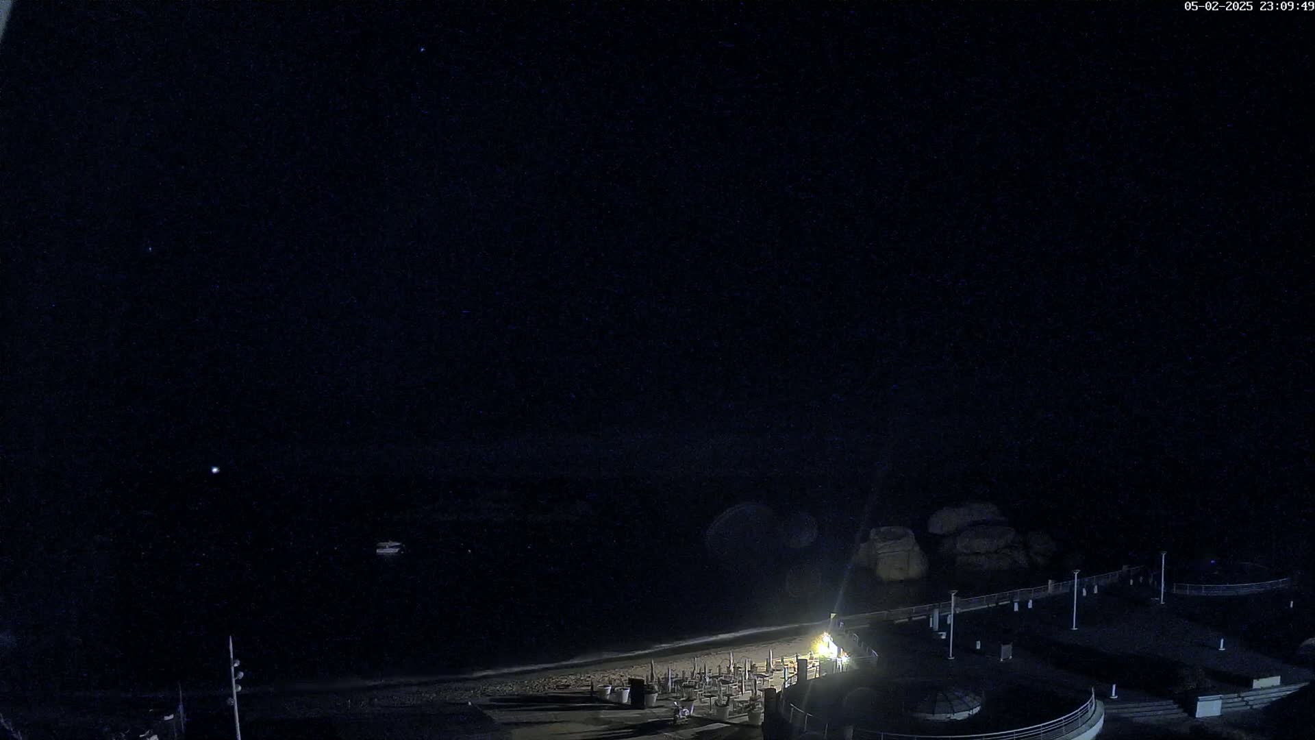 A nighttime, high-angle view shows a dark beach with some lights near the water's edge, under a clear, starry sky.
