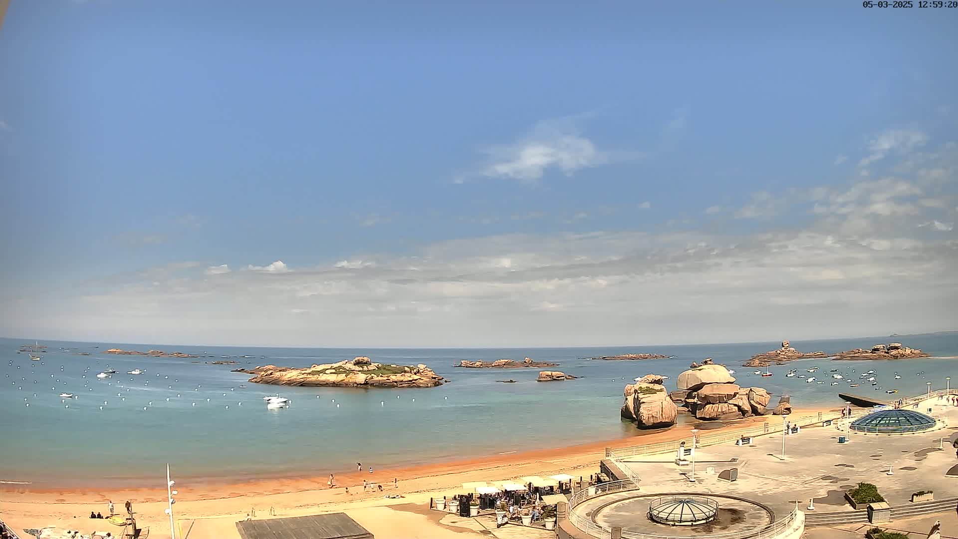 A sandy beach with several small boats and rocky outcroppings in the calm, blue ocean under a mostly sunny sky.