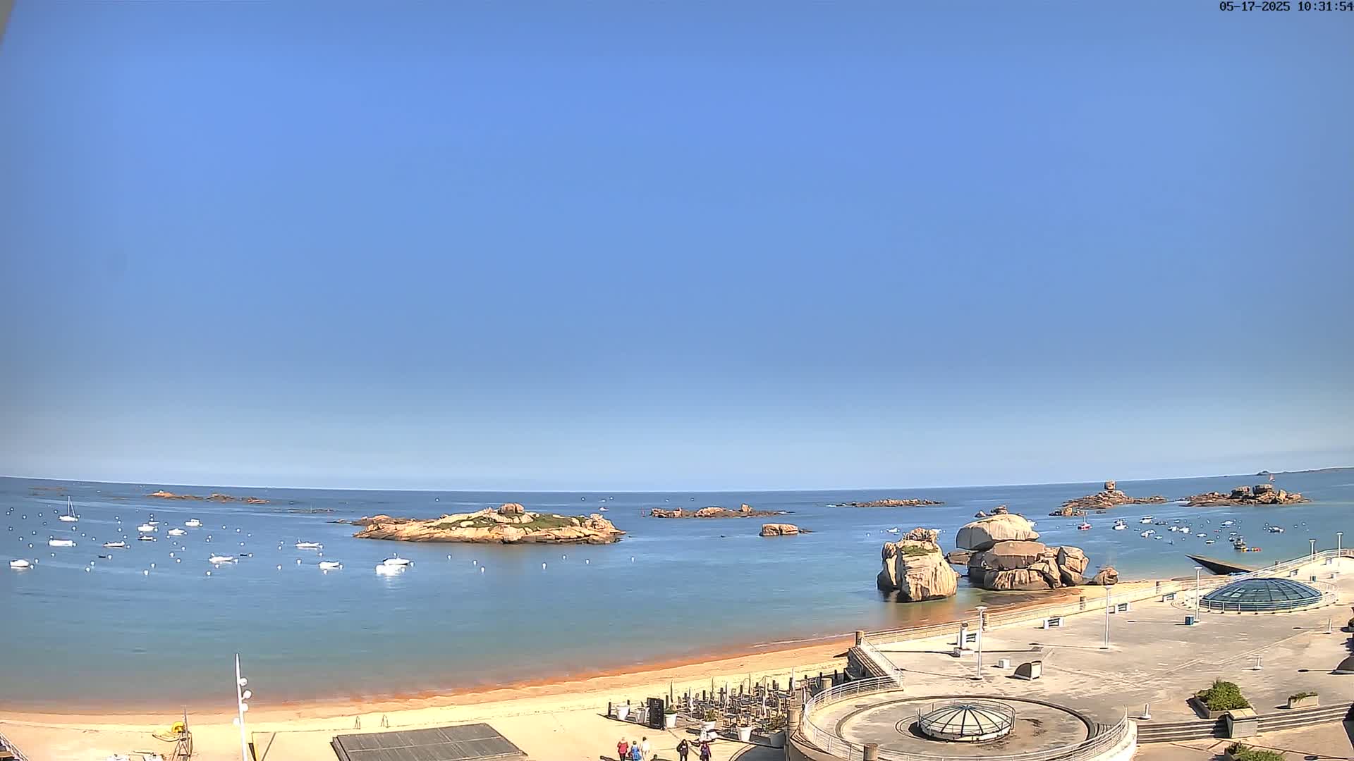 A sandy beach with numerous small boats in calm, clear blue water, under a clear blue sky, and rocky outcrops visible in the ocean.