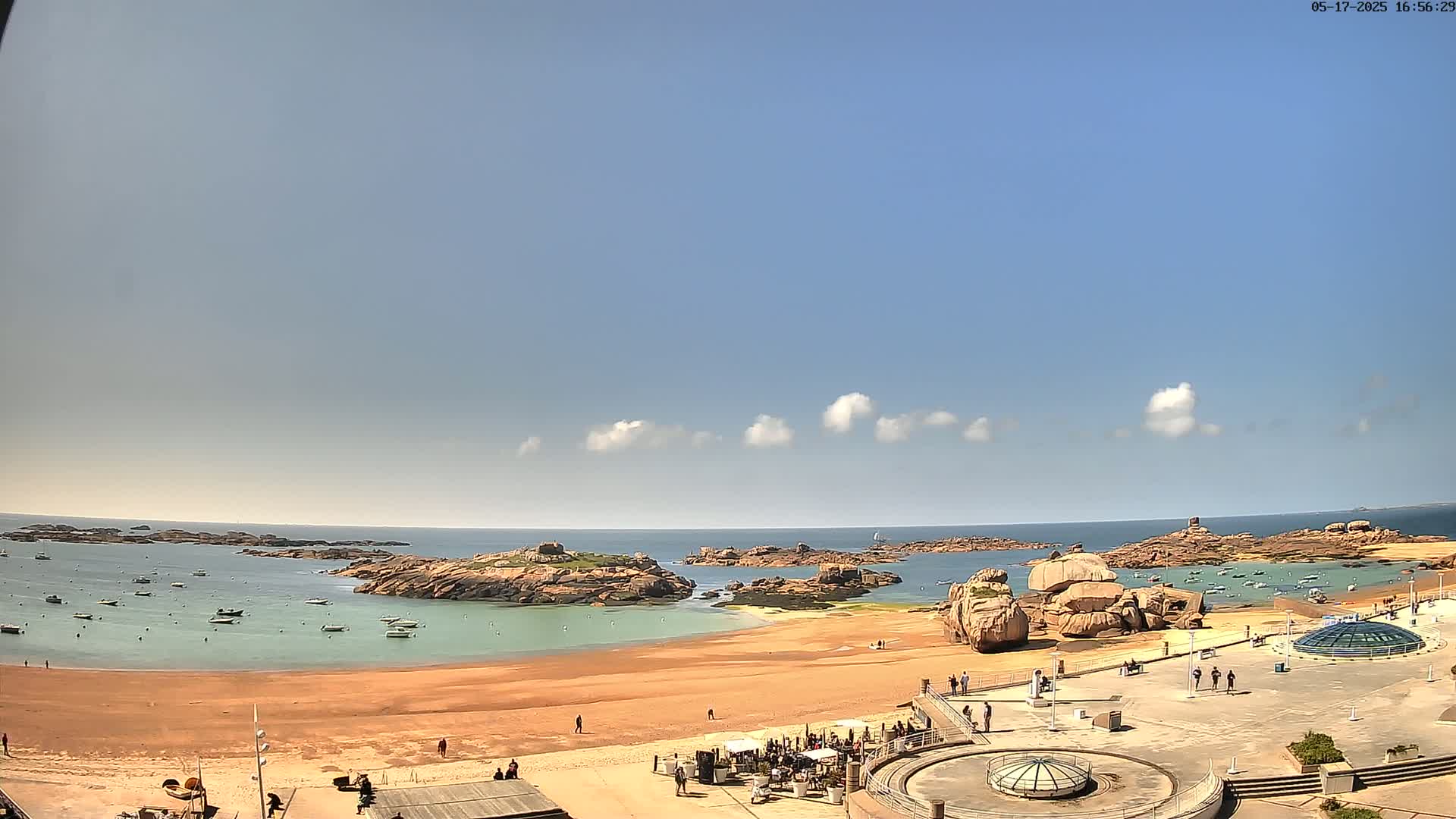 A sandy beach with calm, shallow turquoise water is surrounded by rocky outcrops and a few small boats under a mostly sunny sky with a few scattered clouds.