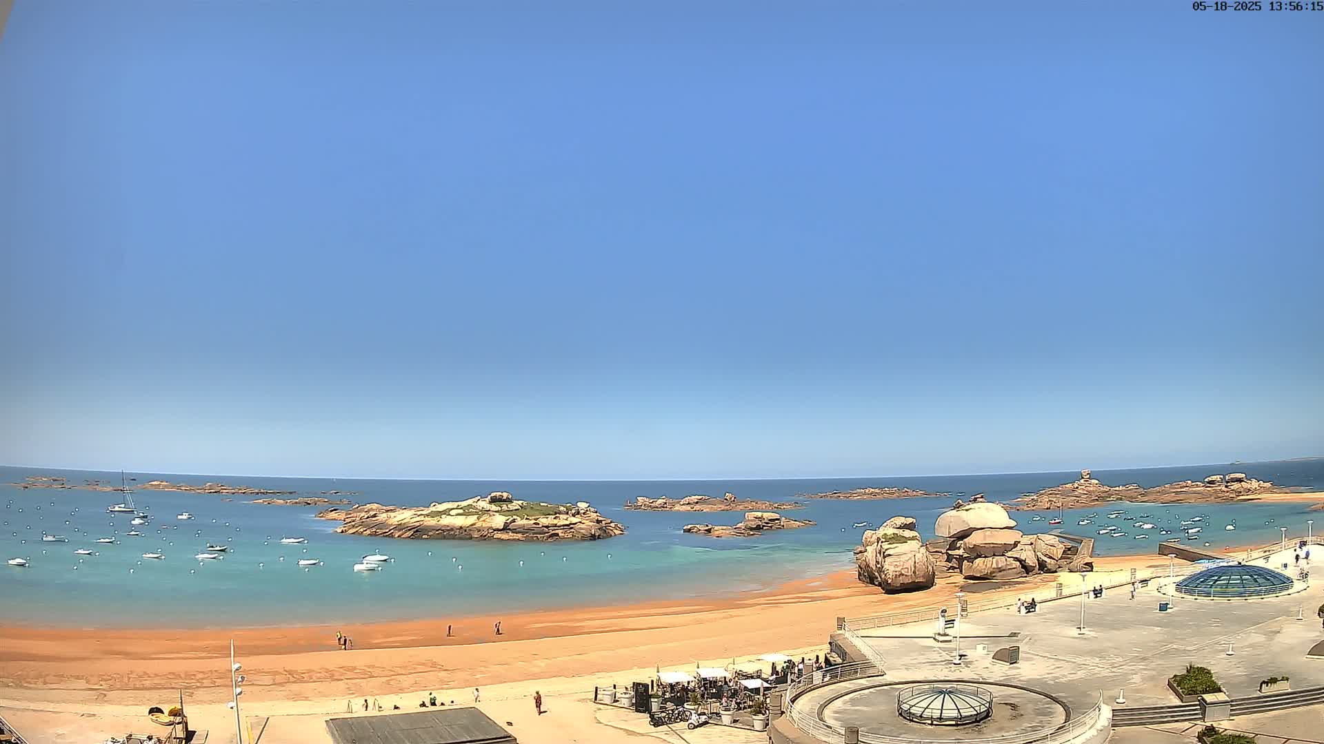 A sandy beach with calm, clear turquoise water, numerous small boats, and rocky outcrops under a clear blue sky.