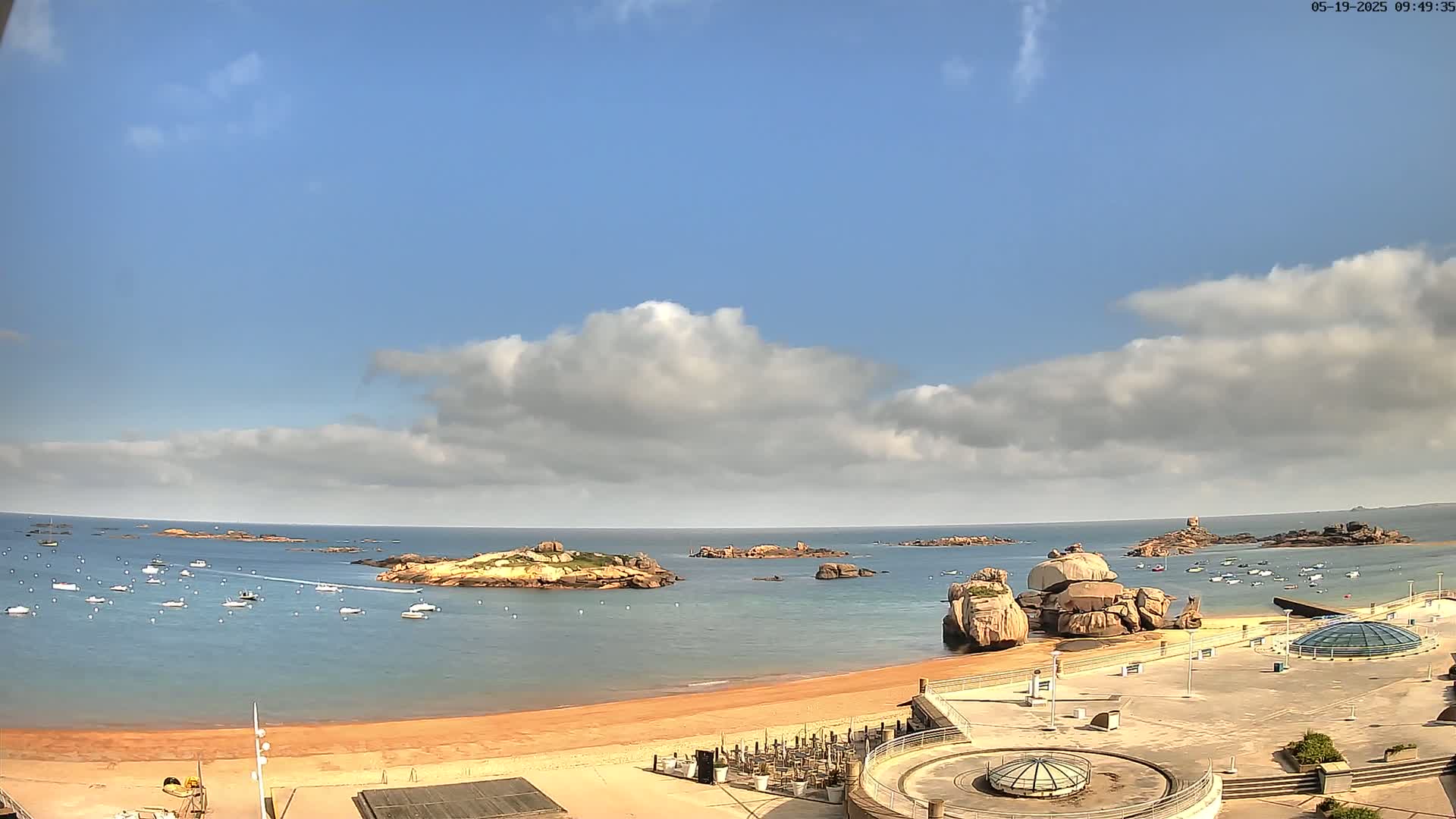 A partly cloudy sunny day reveals a sandy beach, calm ocean water dotted with boats, and rocky outcrops near the shore, all overlooked by a paved plaza.