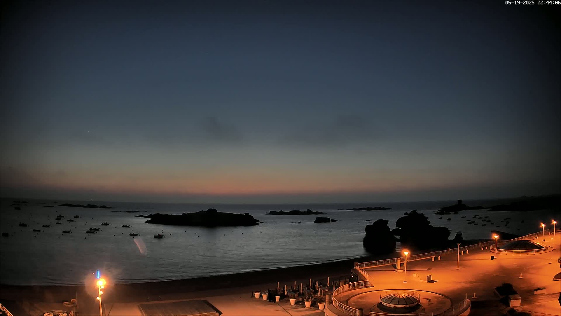 A twilight view of a calm ocean with several small boats and rocky outcrops, a sandy beach, and a paved area with illuminated lampposts.