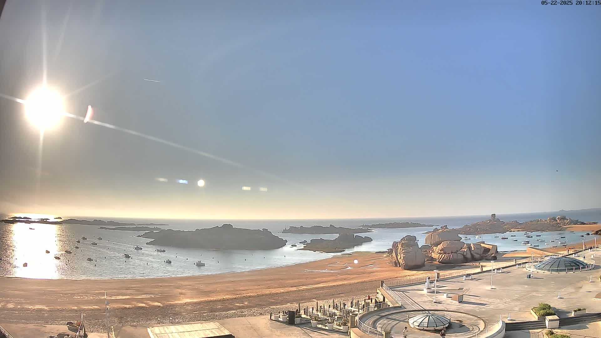 A sandy beach with calm water and several small boats is overlooked by a large flat area with structures and rock formations under a bright, sunny sky.