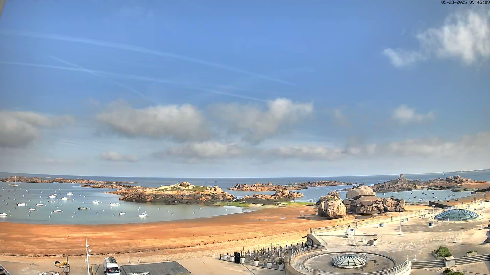A partly cloudy sky overlooks a sandy beach with calm water, numerous small boats, and rocky outcroppings.