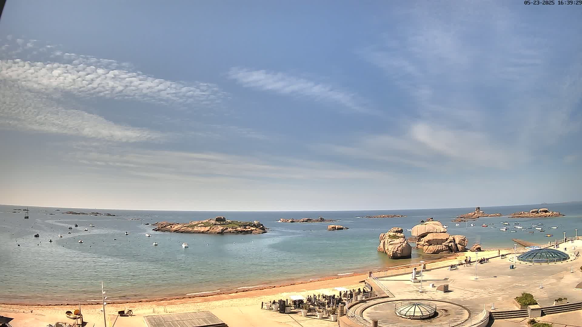 A sandy beach and calm ocean are seen from an elevated vantage point, under a mostly sunny sky with scattered, thin clouds, featuring several rocky outcroppings in the water and numerous small boats.