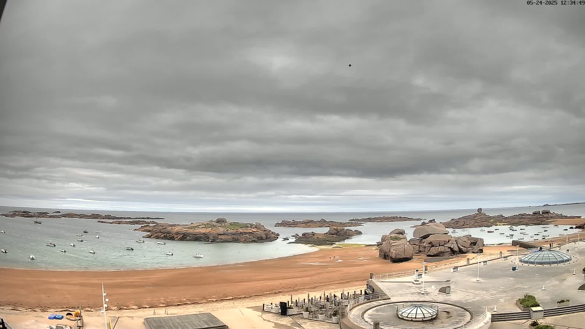 A sandy beach with numerous boats in the calm water, backed by rocky outcrops under an overcast sky.