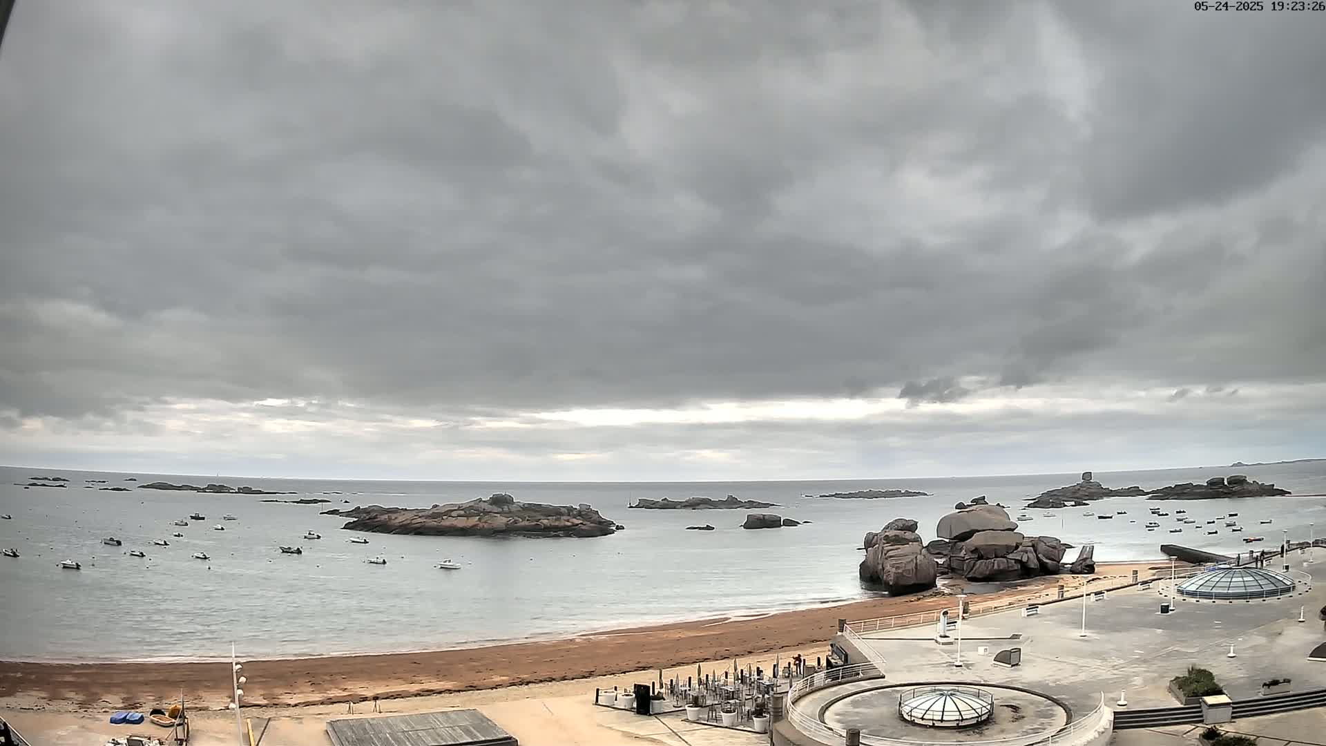 A sandy beach with numerous small boats in the calm water under an overcast sky.