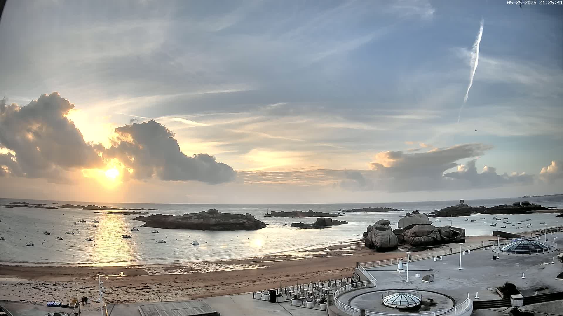 A partly cloudy sunset over a calm bay with numerous small boats and rocky outcroppings, viewed from a seaside building with a flat roof.