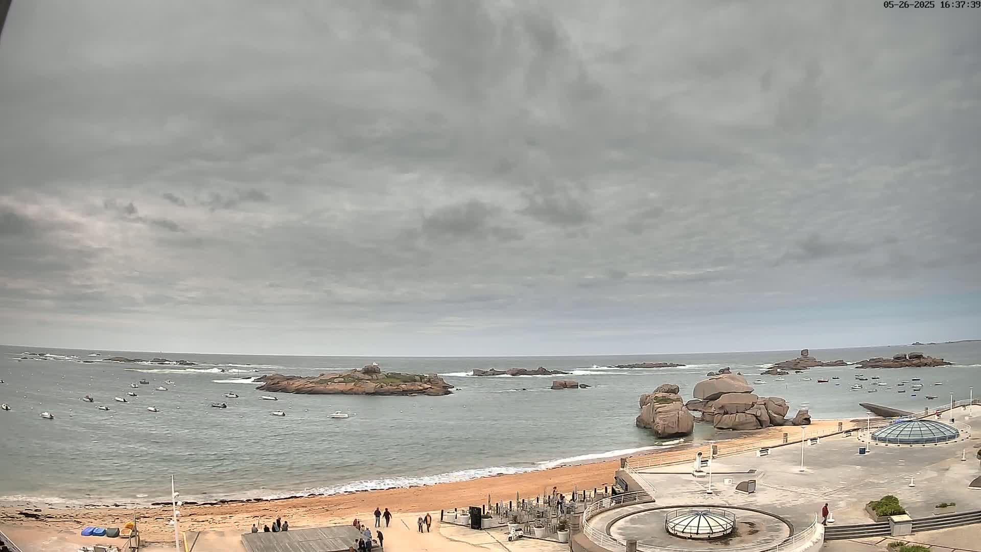 A sandy beach with several small boats in the calm water, overlooked by a paved area, under a cloudy sky.