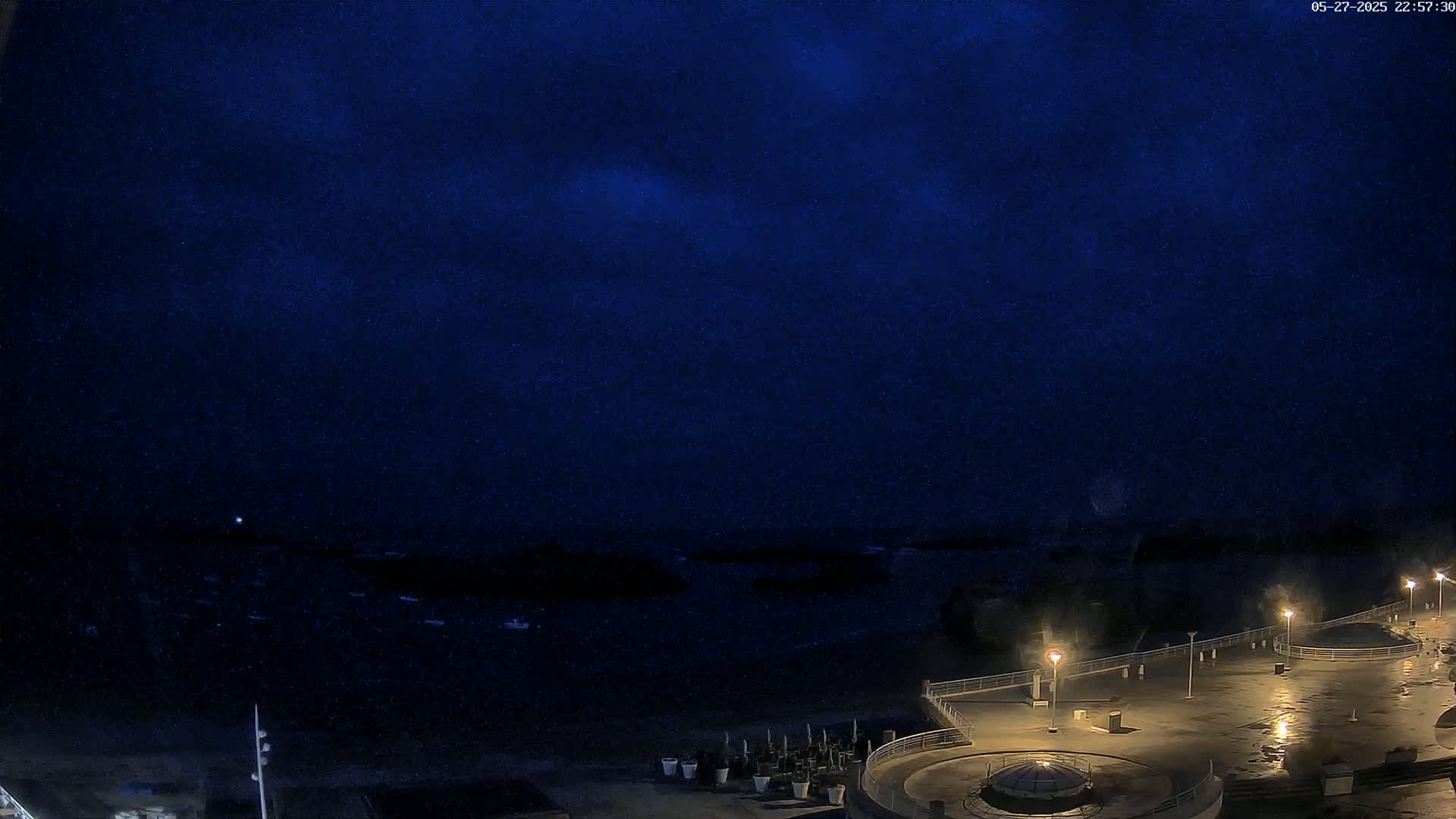 A nighttime, dark blue sky overlooks a dimly lit, empty waterfront promenade with a few lights and potted plants.