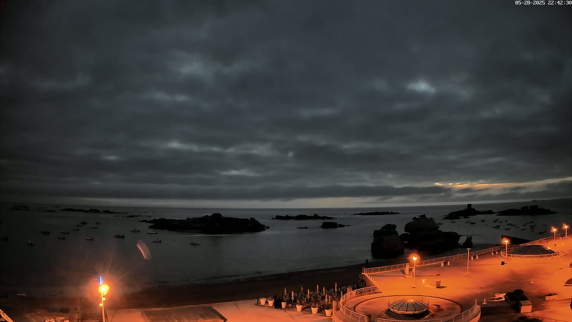 A dark, cloudy night scene shows a rocky coastline with a calm body of water, several small boats, and a paved area with illuminated lamps and structures.