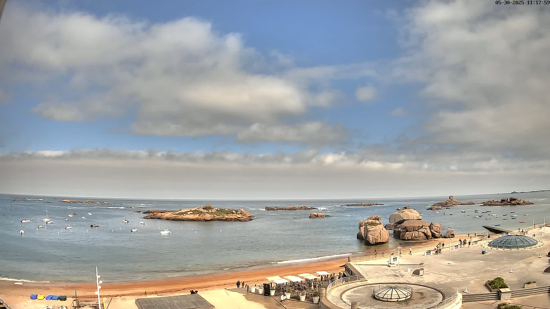 A partly cloudy sky overlooks a sandy beach with numerous small boats in the calm water near rocky outcrops.