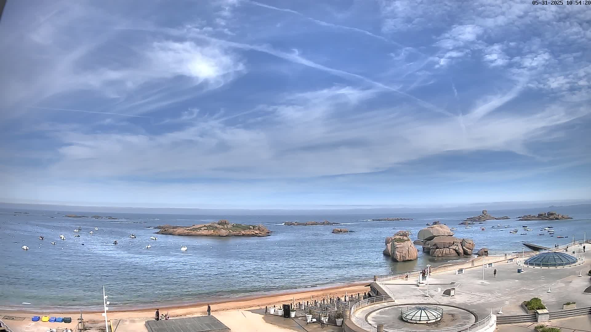 A mostly sunny day shows a calm bay with numerous small boats, rocky outcroppings, and a sandy beach with a paved area containing several structures.