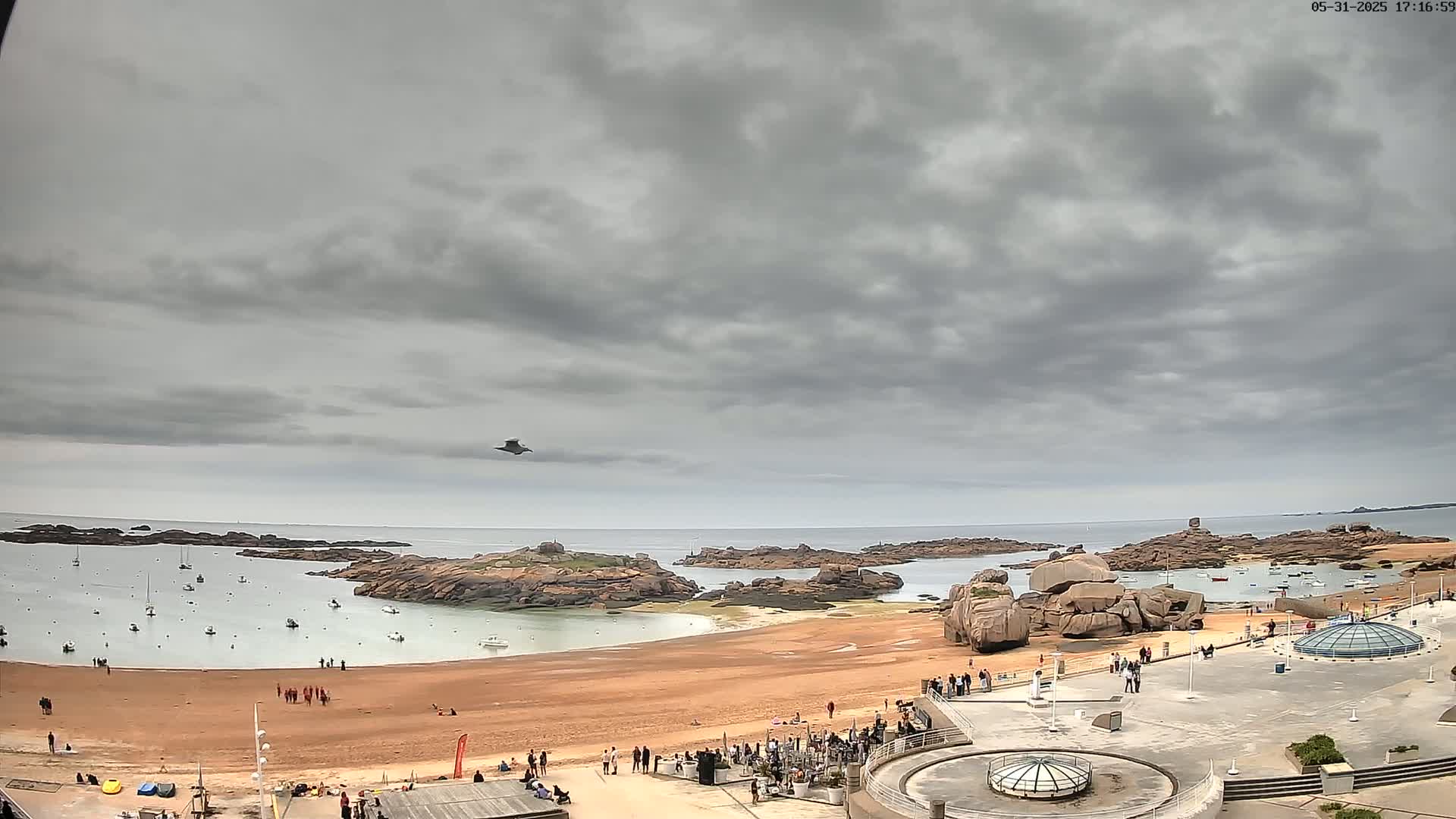 A sandy beach with scattered people and boats in the calm water is backed by rocky outcrops under an overcast sky, a bird flying overhead.