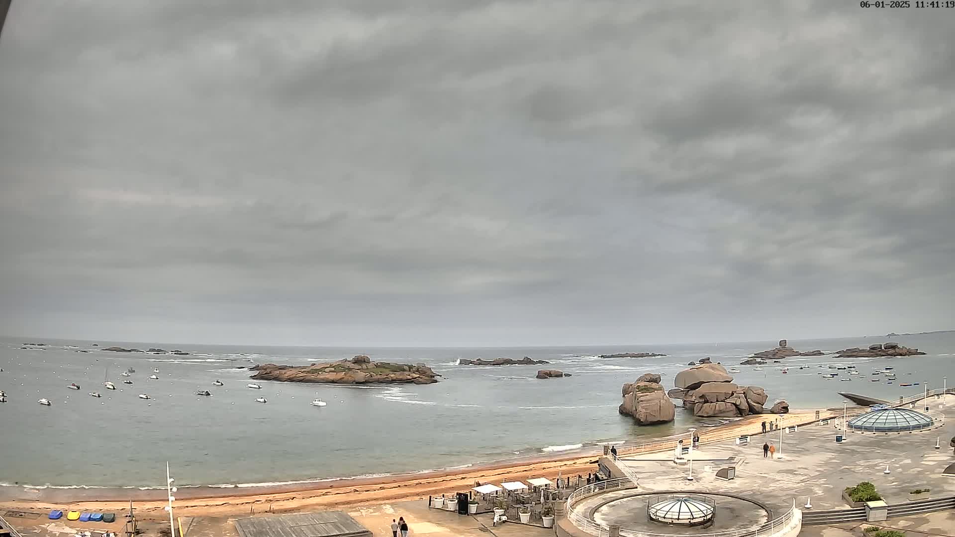 A sandy beach with several boats in the calm water and large rocks jutting out of the ocean under an overcast sky.