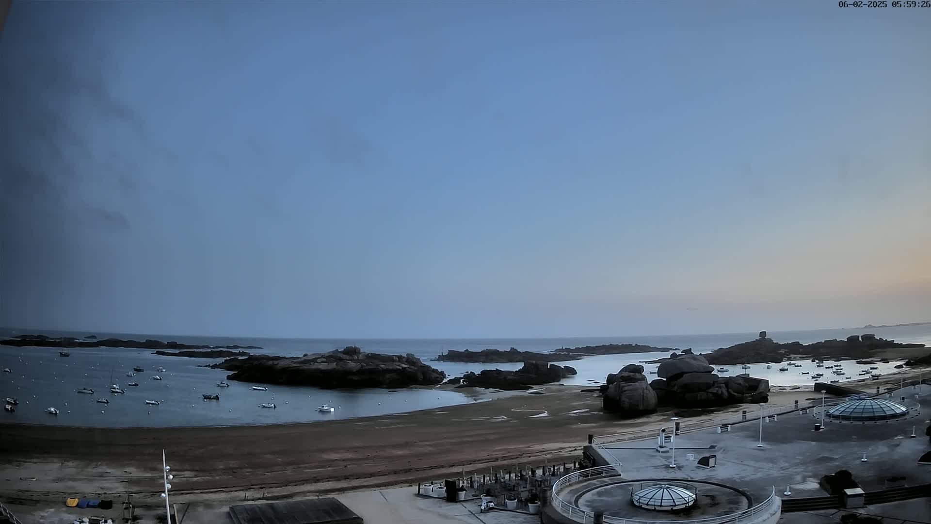 A calm, clear morning reveals a rocky coastline with numerous small boats in a bay, adjacent to a sandy beach and a paved area with several structures.