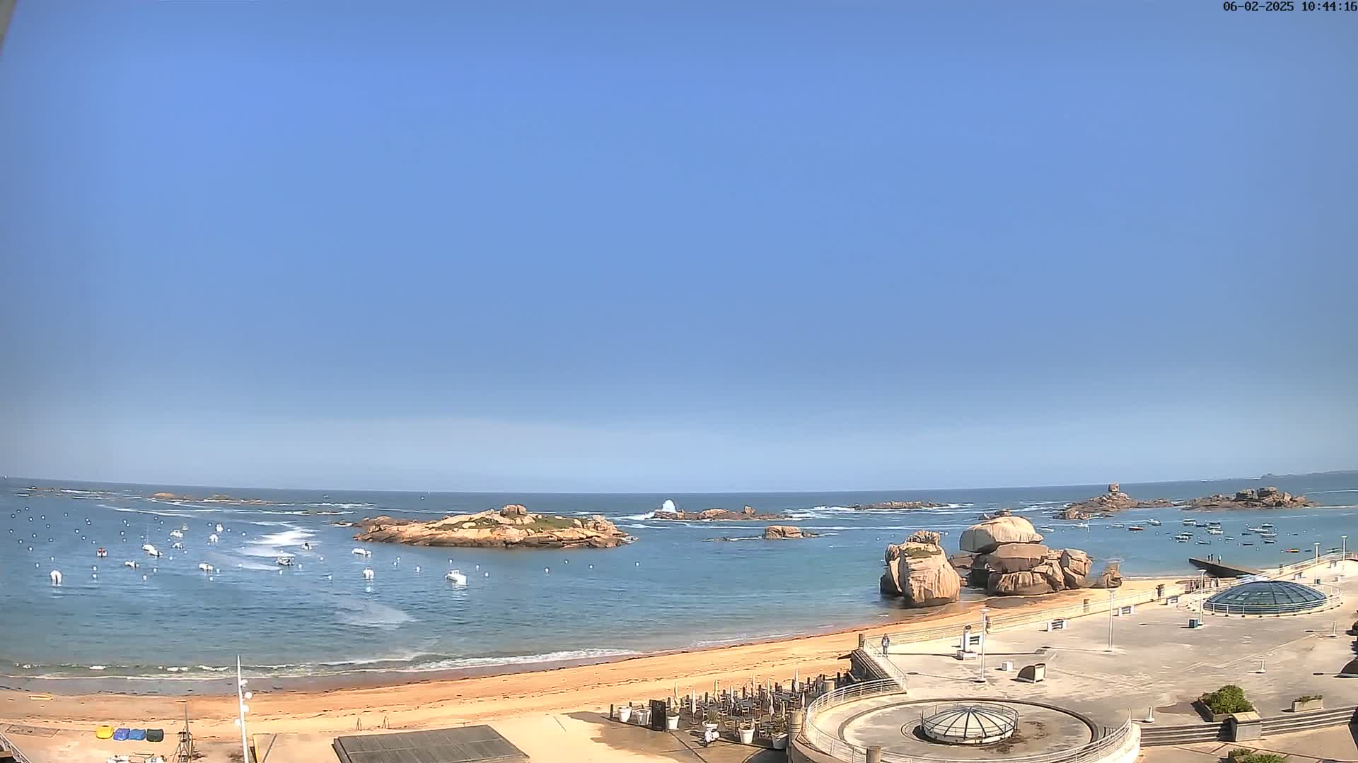 A sandy beach with numerous small boats in the water, rocky outcroppings, and a clear blue sky under sunny conditions.
