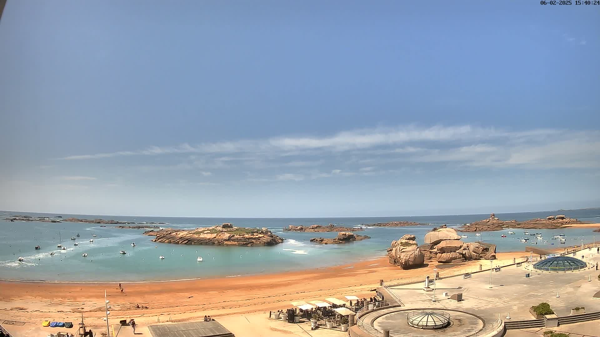 A sandy beach with calm, teal water, several rocky islands, and a few boats is visible under a mostly clear, sunny sky with some thin clouds.