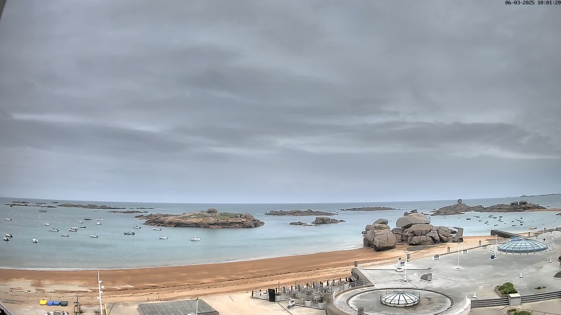 A sandy beach with several boats in the calm water, overlooked by rocky outcroppings under a cloudy sky.