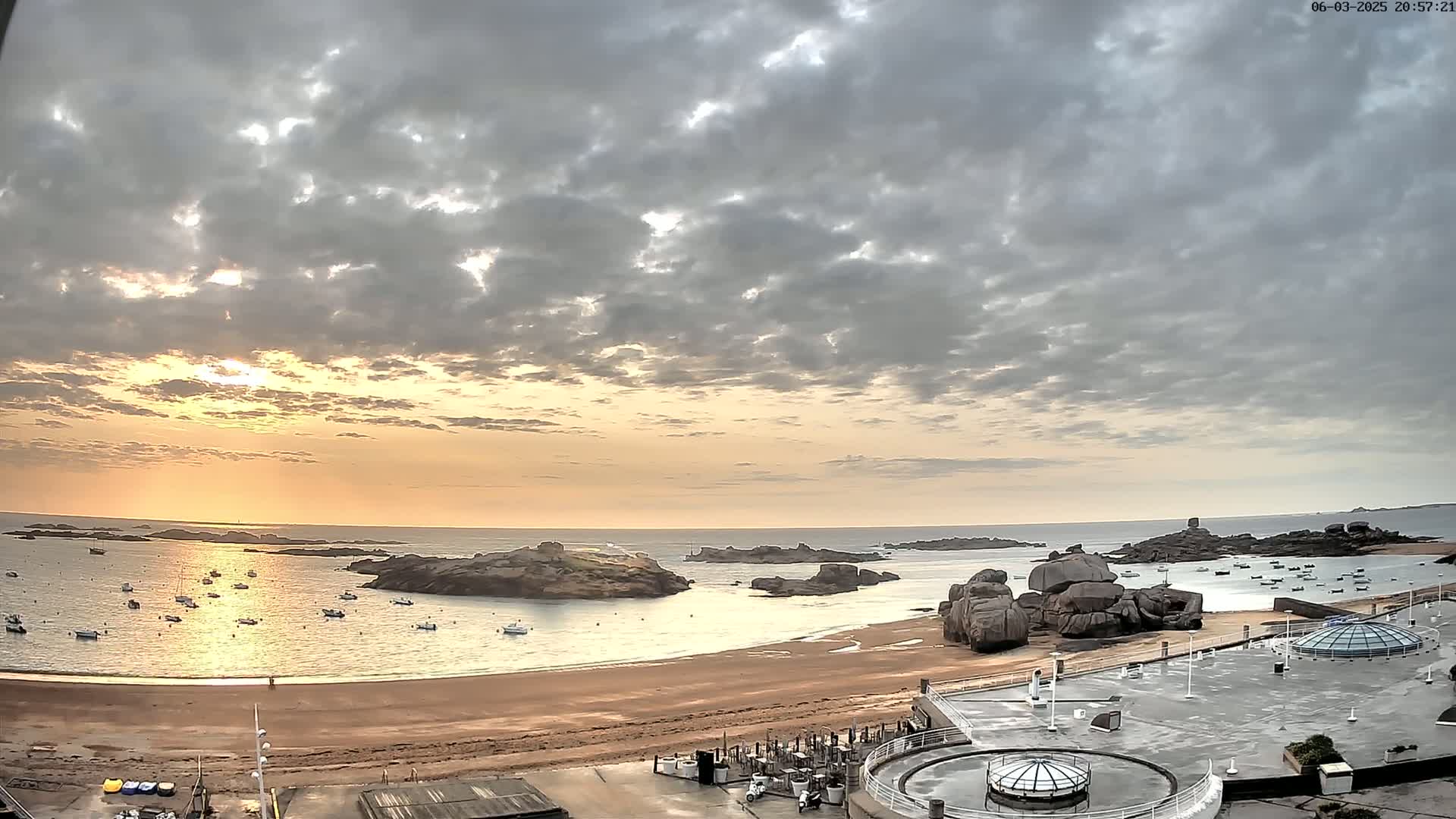 A sandy beach with calm water and numerous boats is visible at sunset under a partly cloudy sky.