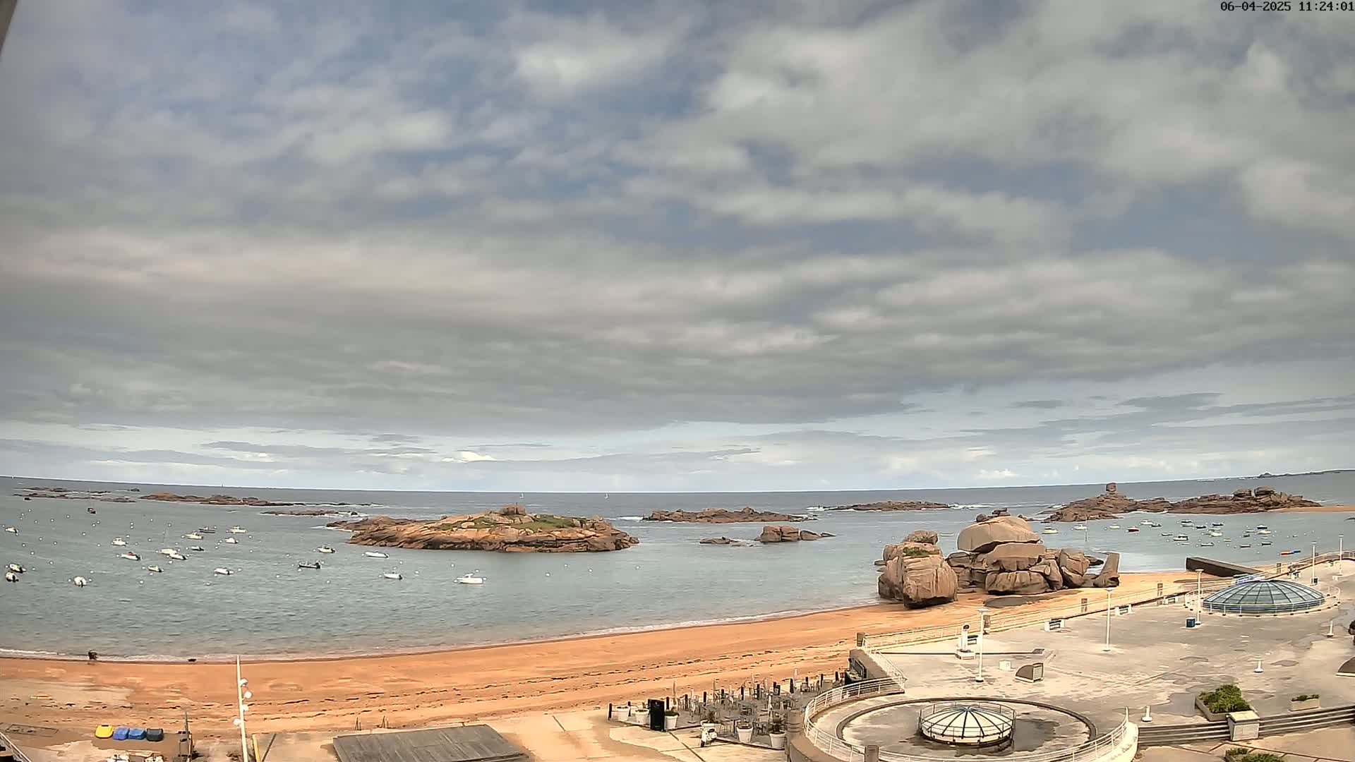 A sandy beach with numerous small boats in the calm water, rocky outcrops in the sea, and an overcast sky.