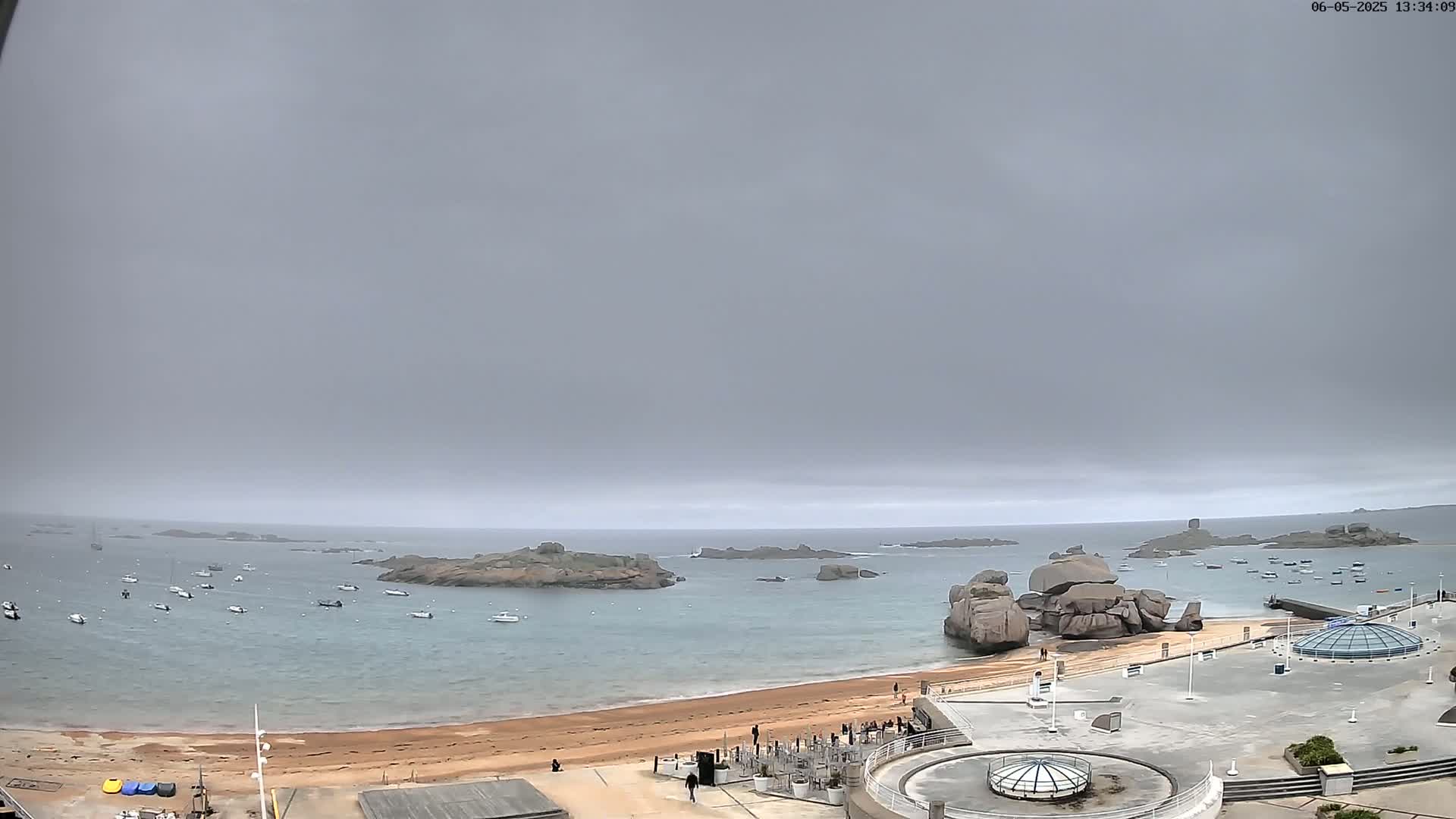 A sandy beach with several boats in the water, rocky outcroppings, and a few people are visible under an overcast sky.