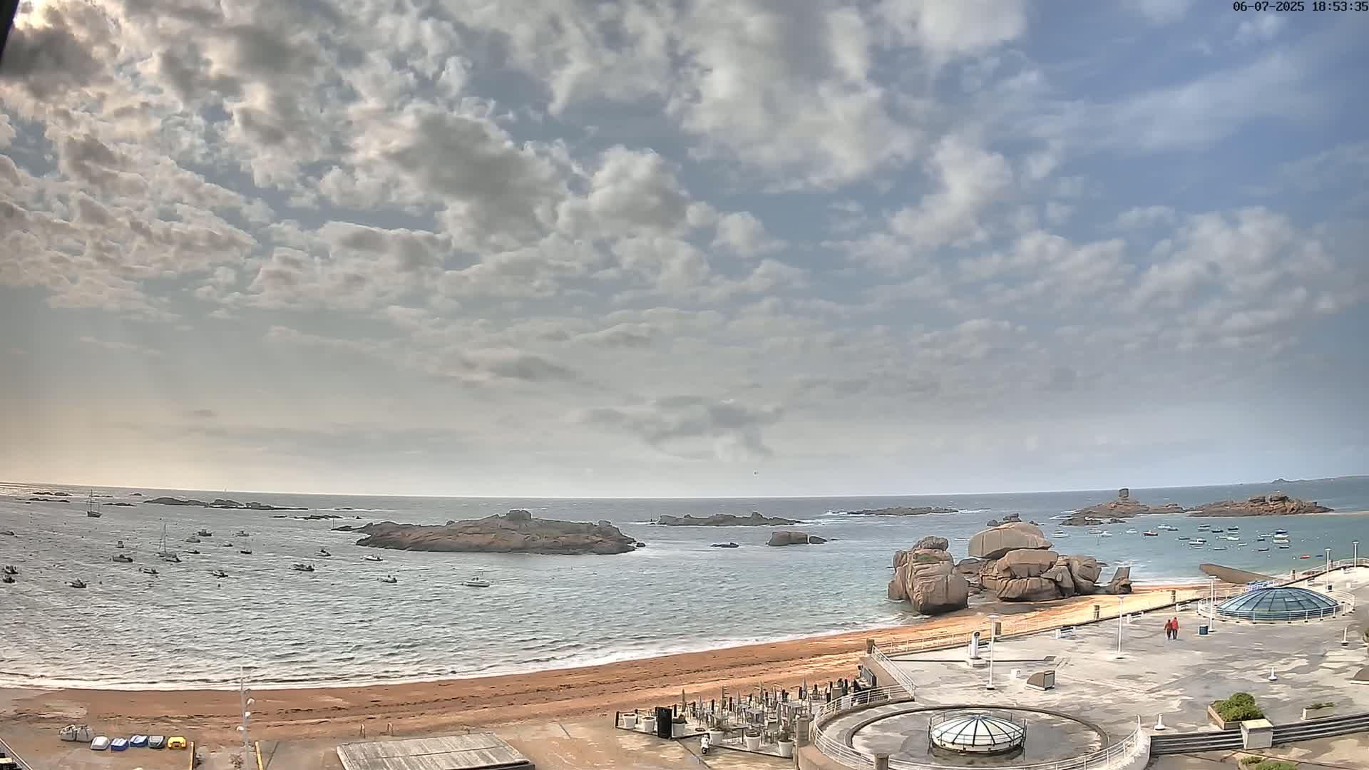 A sandy beach with several boats in the water, rocky outcroppings, and a partly cloudy sky.