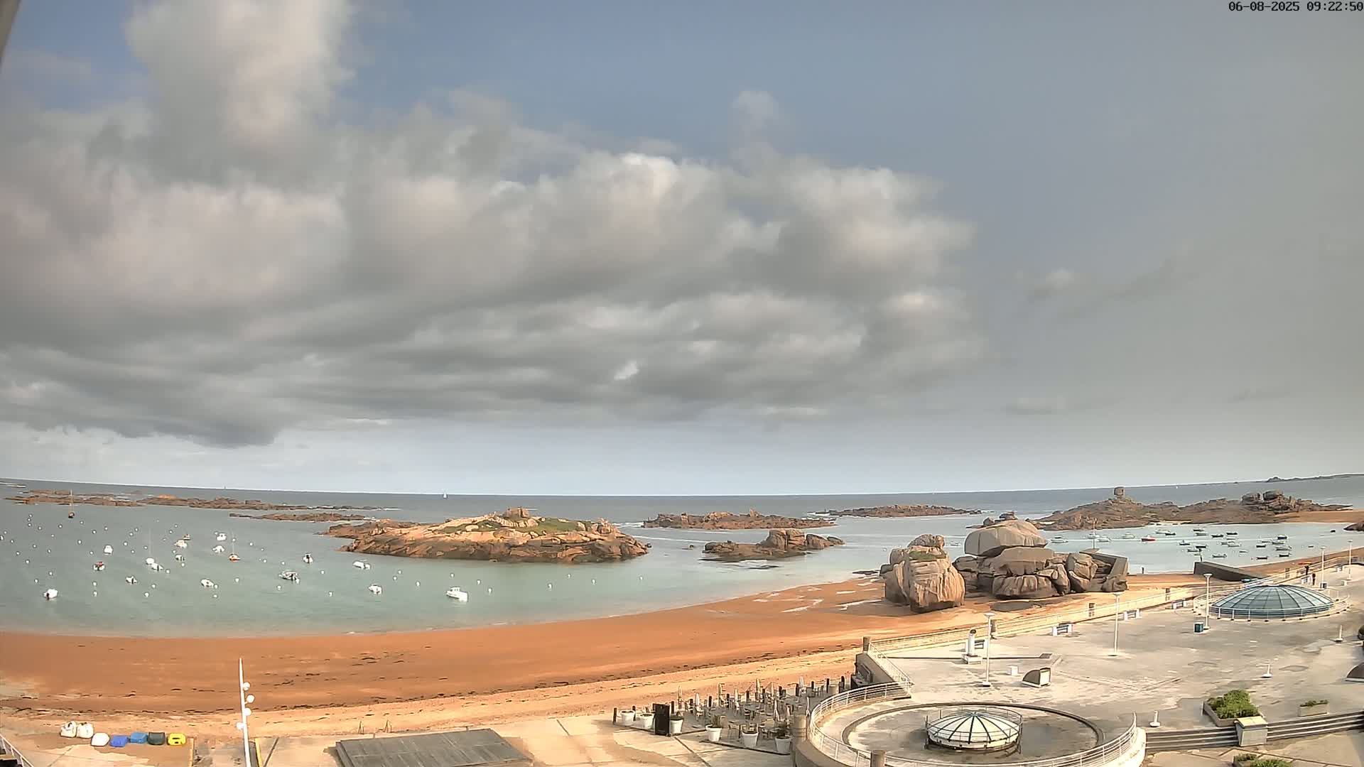 A sandy beach with numerous small boats in the calm water, overlooked by large rocks and a partly cloudy sky.