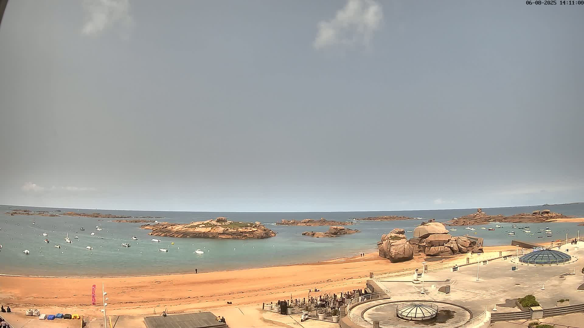 A sandy beach with numerous boats in the calm, light-grey-skied water, surrounded by rocky outcrops.