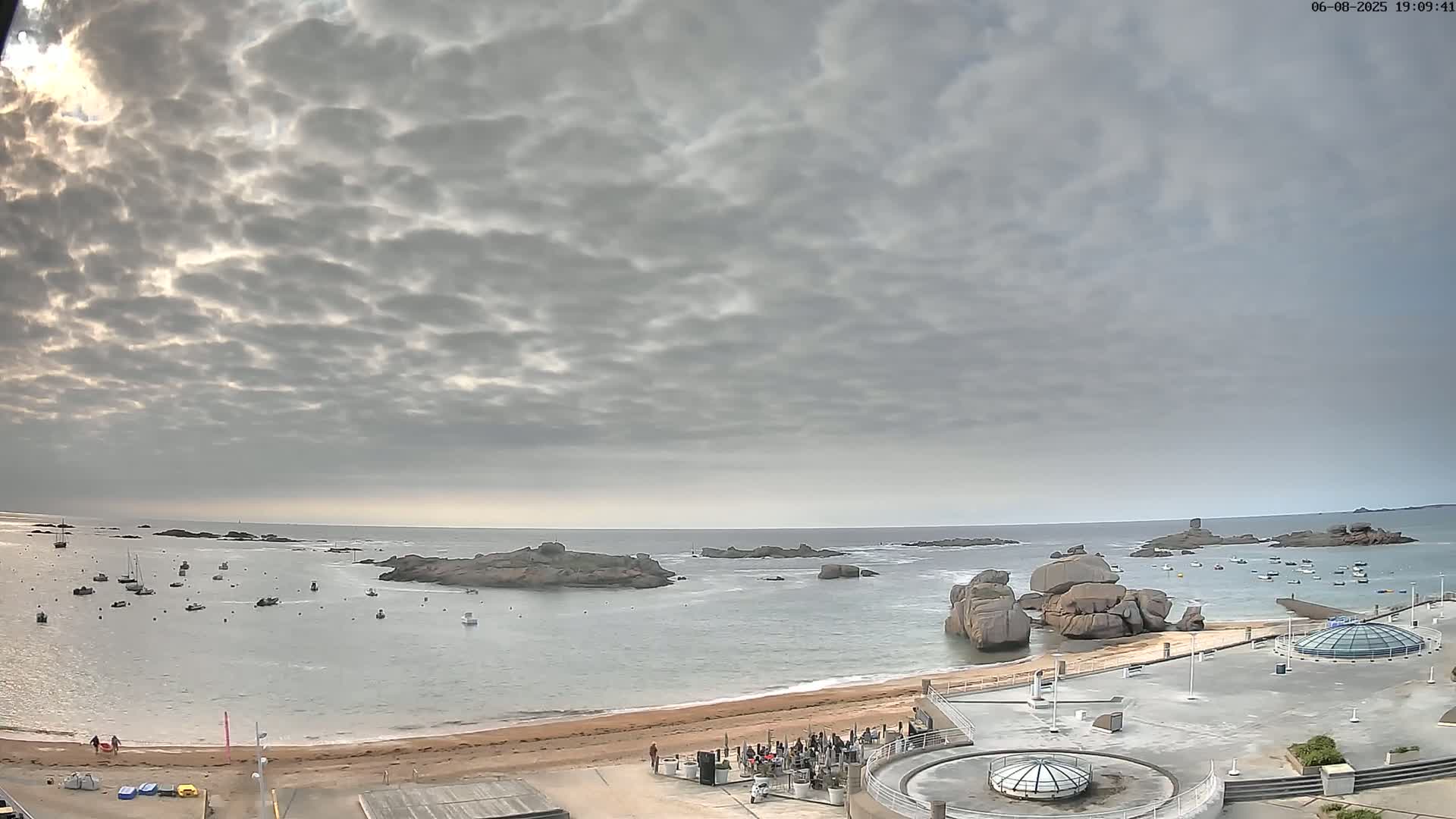 A sandy beach with several boats in the calm water, large rocky formations offshore, and an overcast sky.