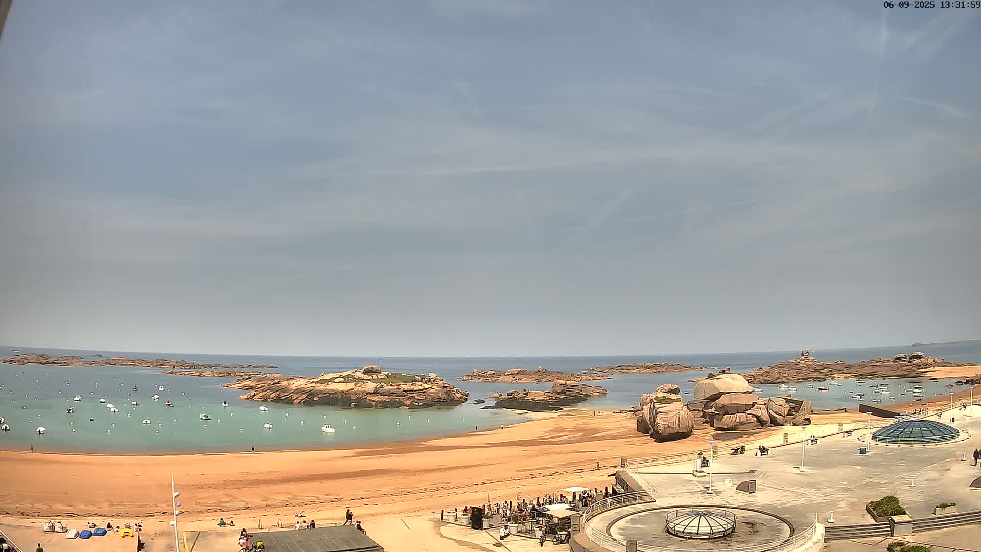 A sandy beach with calm, light-blue water, rocky outcroppings, and a few boats is seen under a mostly clear, light-blue sky.