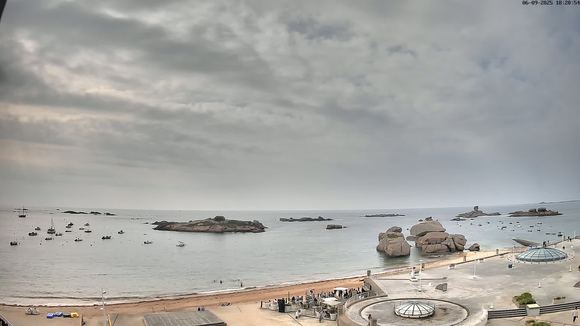 A sandy beach with several boats in the calm water near rocky outcrops under an overcast sky.