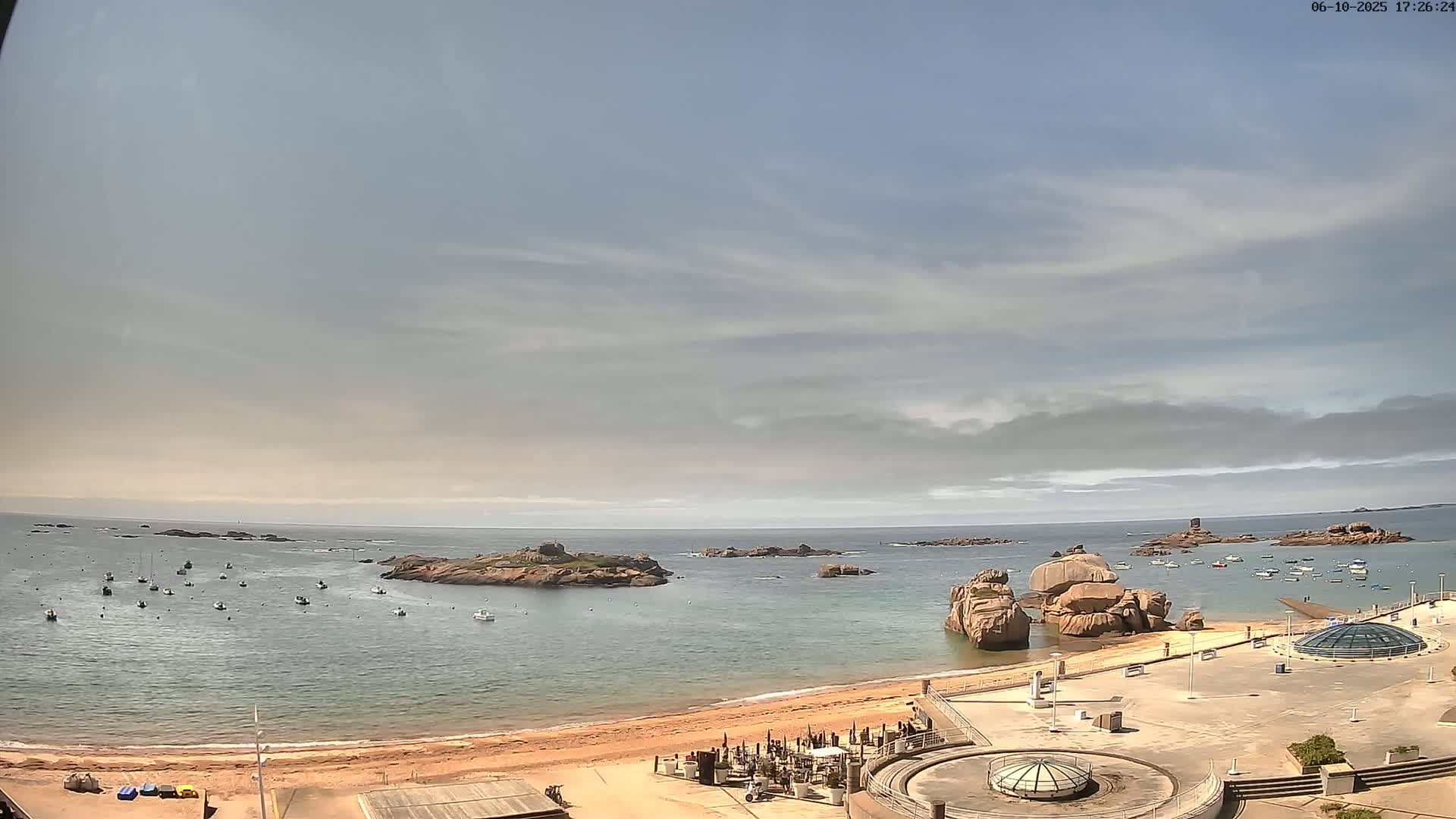 A sandy beach with several boats in the calm water, overlooking rocky islands under a mostly cloudy sky.