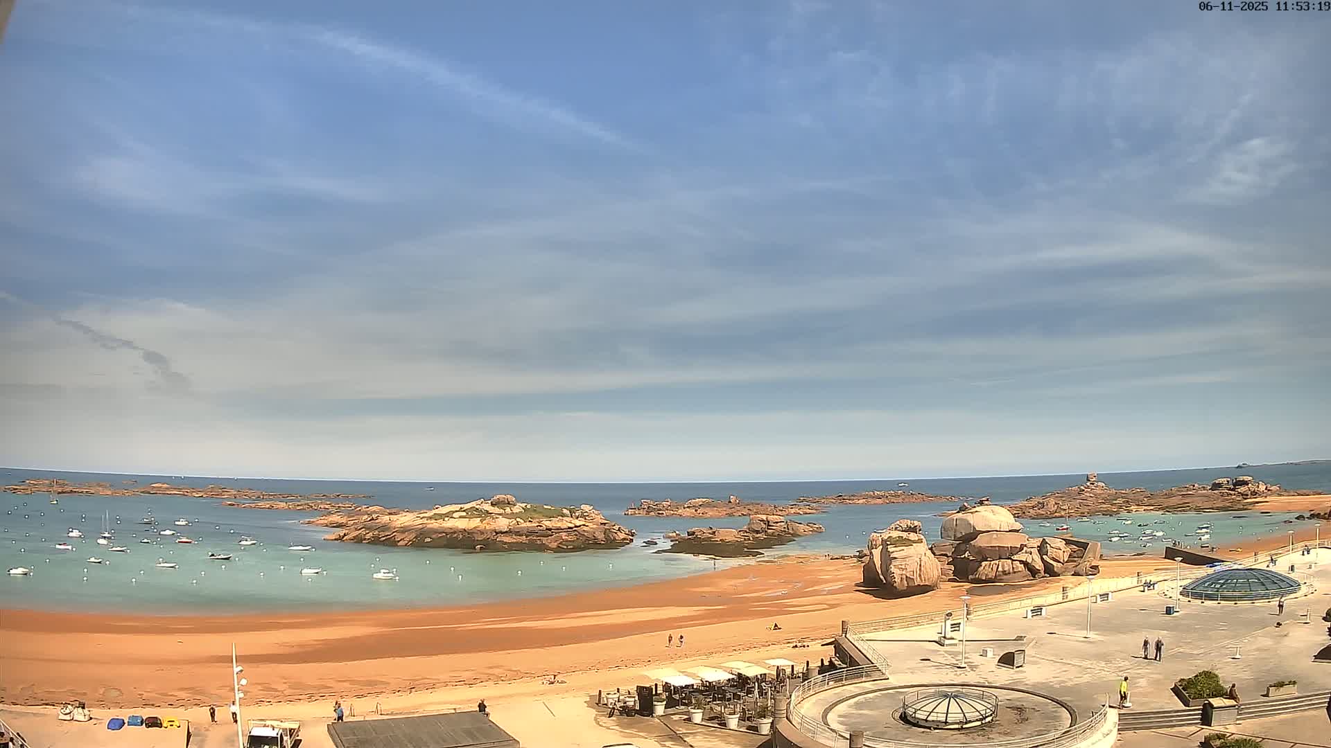 A sandy beach with calm, light-green water and several boats is surrounded by rocky outcrops under a mostly sunny sky with a few scattered clouds.