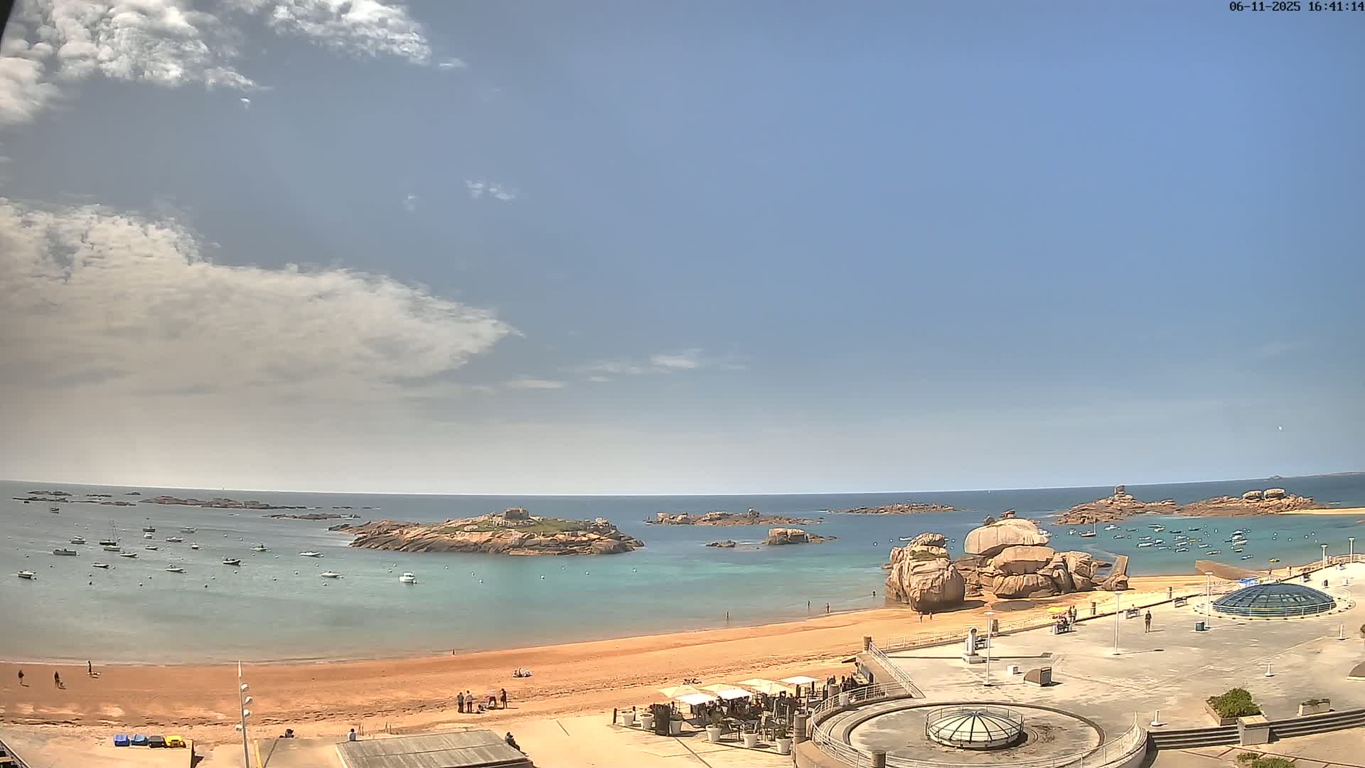 A partly cloudy day reveals a sandy beach with several small boats in the calm, teal water near rocky outcrops and a paved plaza with a few people.