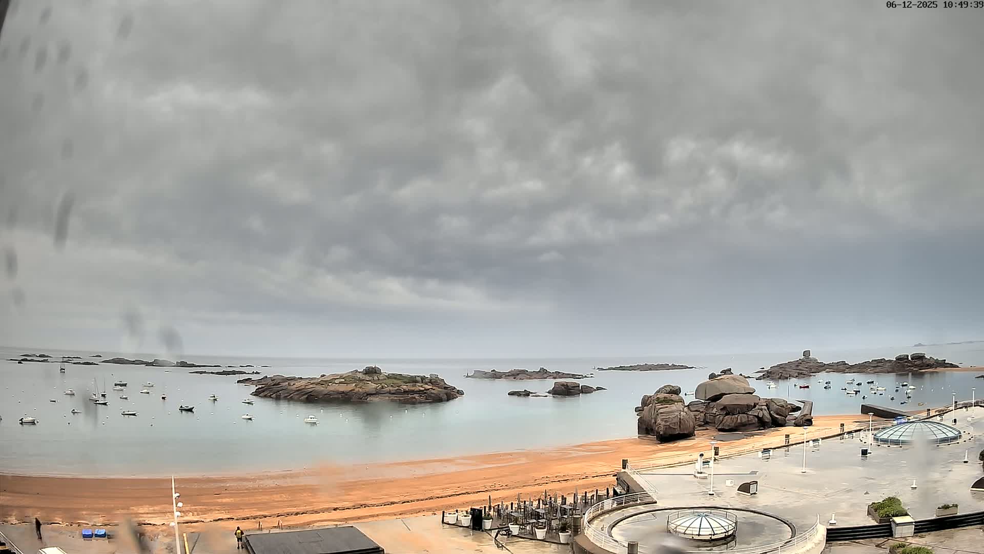 A sandy beach with numerous small boats in the calm water, surrounded by rocky outcrops under an overcast sky.