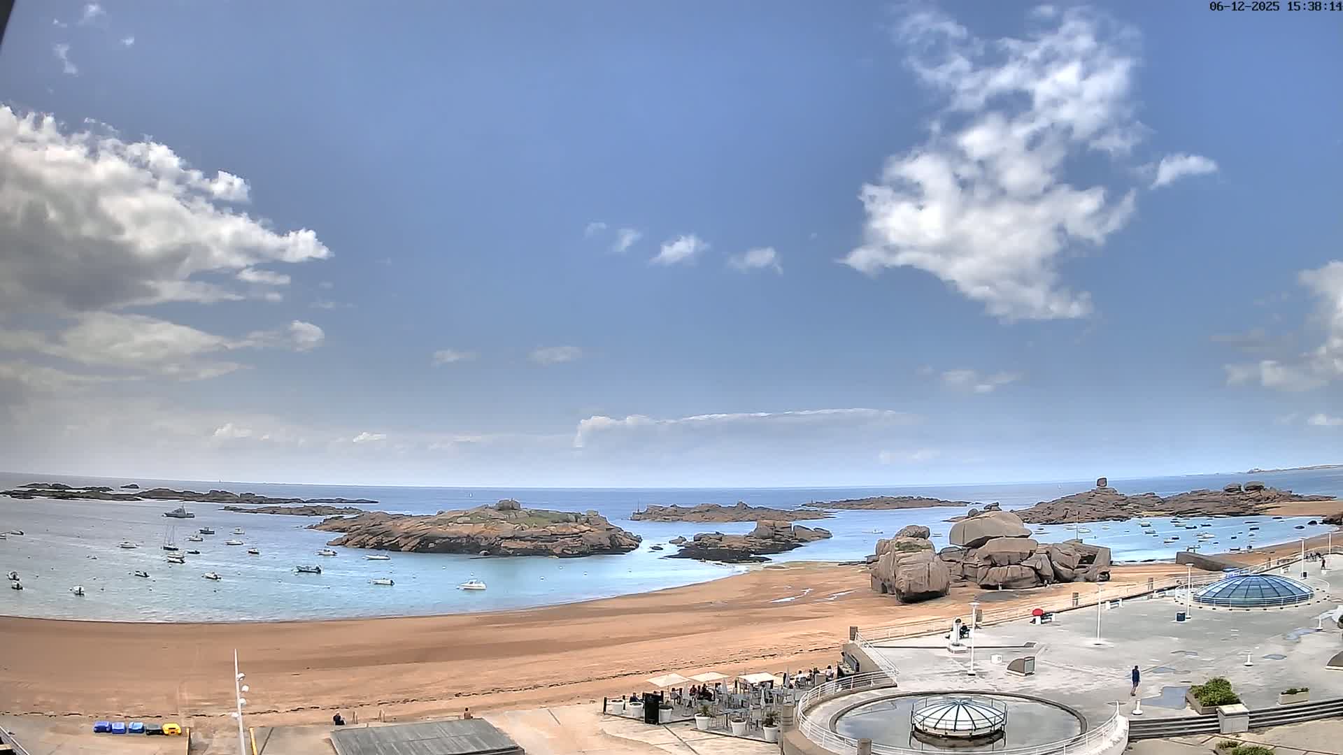 A sandy beach with calm water and numerous boats is surrounded by rocky outcrops under a mostly sunny sky with scattered clouds.