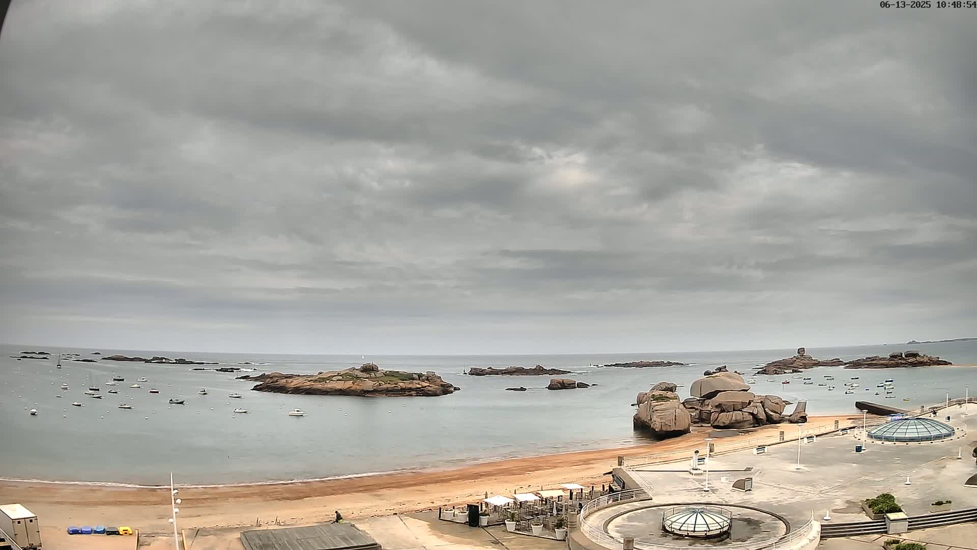 A sandy beach with several boats in the calm water, rocky islands offshore, and an overcast sky.