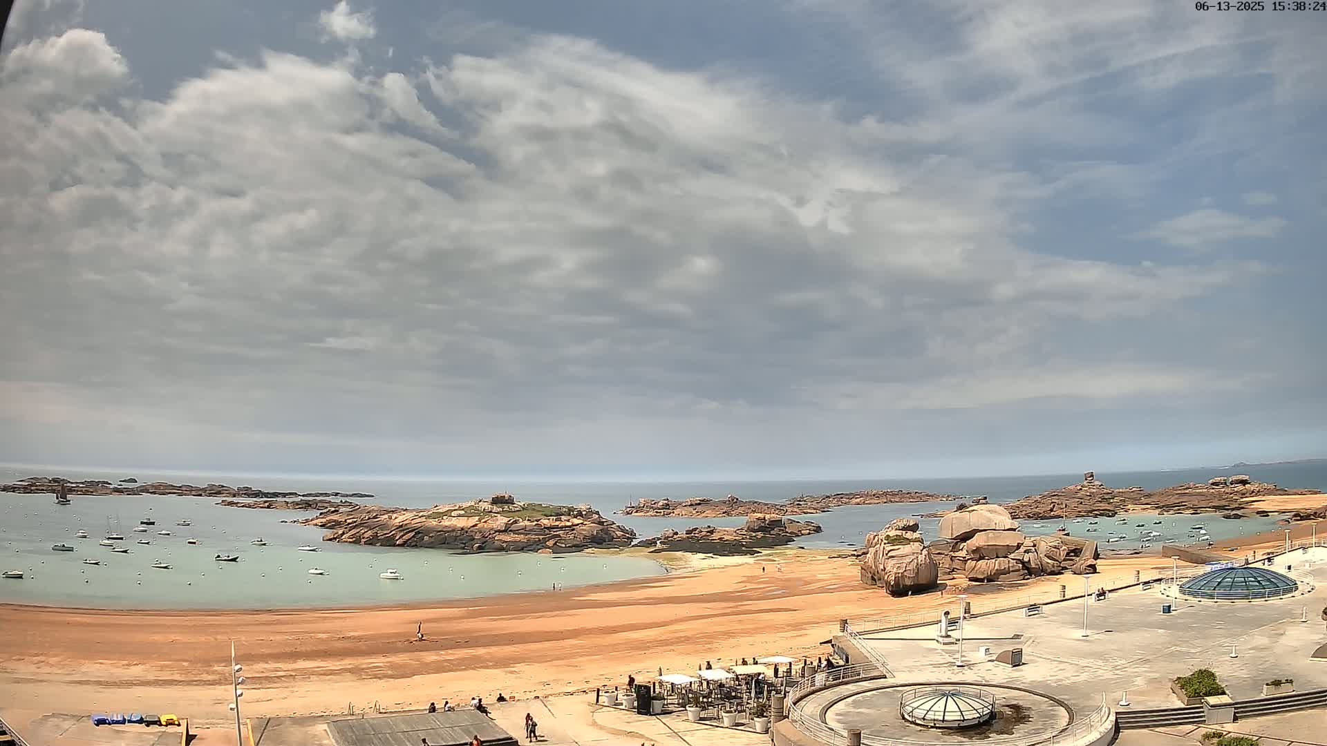 A partly cloudy day reveals a sandy beach, calm water dotted with small boats, and rocky outcrops extending into the sea.