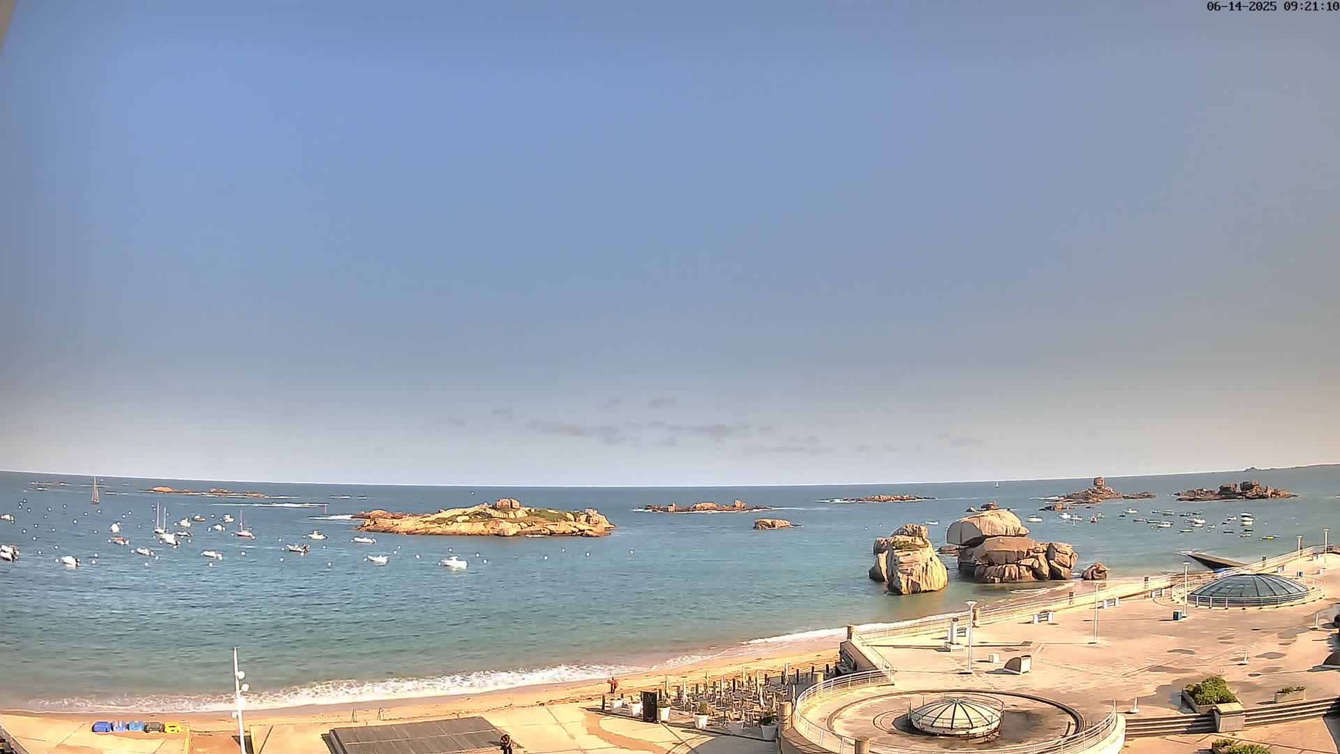 A sandy beach with several boats in the calm ocean, rocky outcrops, and a clear blue sky.